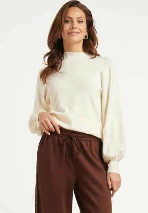 Vrouw met een witte longsleeve met pofmouwen en een bruine broek met koord, staand en glimlachend tegen een effen achtergrond.