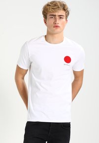 Fehér pamut póló kerek piros grafikával és japán szöveggel a bal mellrészen. Rövid ujjú és kerek nyakú.