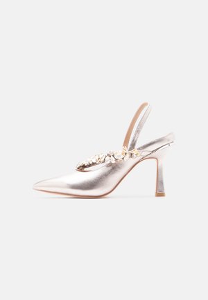 Escarpin à bout pointu argenté avec bride arrière et ornements en pierres précieuses transparentes et dorées sur fond blanc.