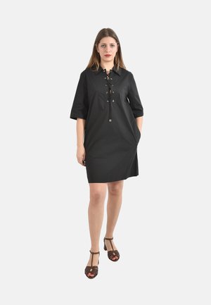 Donna che indossa un vestito nero al ginocchio con chiusura frontale a lacci e maniche a tre quarti, abbinato a sandali con tacco alto marrone scuro con cinturino a T.