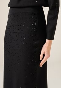 Gonna nera in maglia con paillettes quadrate sparse su tutto il tessuto. Il tessuto è liscio con una silhouette aderente, mostrata accanto a una mano.