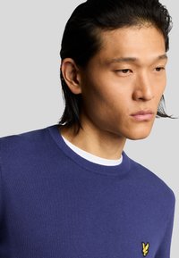 Felpa a maglia viola con scollatura rotonda e un sottile logo giallo sul lato sinistro del petto. La texture è liscia e la vestibilità è rilassata.
