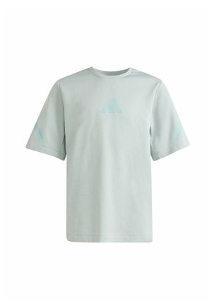 Niño de pie con una mano en el bolsillo y sosteniendo una gorra gris, vistiendo una camiseta gris clara, pantalones deportivos negros con rayas blancas y zapatillas negras.