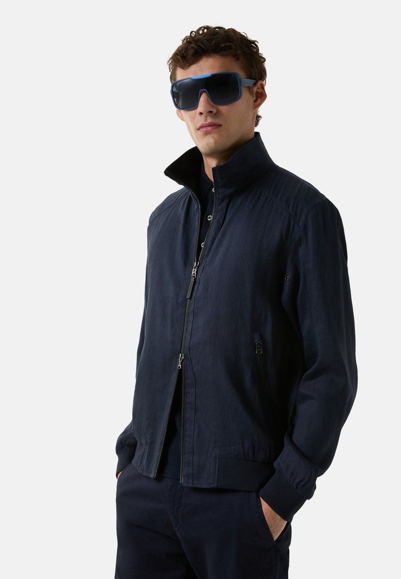 Veste de bombardier marine avec un col montant, fermeture éclair et poches latérales. Le tissu a une texture lisse et un éclat subtil. Portée avec de grandes lunettes de soleil.