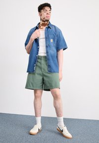 Camisa de manga corta de denim en azul claro, pantalones cortos de algodón verdes y zapatillas blancas con detalles en azul marino. Modelo de pie contra un fondo liso.