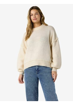 Jeune femme aux longs cheveux blonds portant un pull en tricot crème à manches bouffantes et un jean bleu, debout devant un fond blanc.