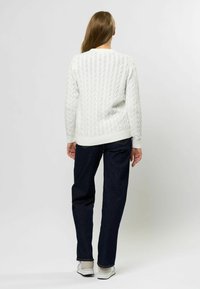Fehér cable-knit pulóver hosszú ujjakkal, bordázott mandzsettával és szegéllyel. Sötét farmernadrággal és világos színű lábbelivel párosítva.