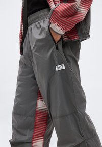 Άτομο που φοράει γκρι παντελόνι φόρμας EA7 Emporio Armani με φερμουάρ στην τσέπη και μανίκι μπουφάν με κόκκινη ρίγα ορατό.