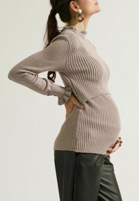 Zwangere vrouw in een beige geribde trui en zwarte broek houdt haar buik met één hand vast en steunt met de andere op haar rug, met grote ronde oorbellen.