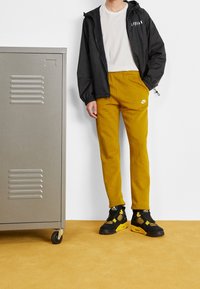 Černá bunda na zip, bílé tričko, hořčicově žluté tepláky s logem Nike a černé tenisky se žlutými akcenty vedle šedé skříňky.