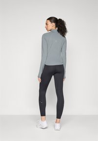 Maglia grigia a maniche lunghe con colletto alla coreana, abbinata a leggings neri. Caratteristiche includono un fit aderente, una texture liscia e dettagli del logo discreti.