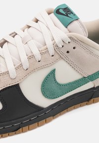 Gros plan d'une paire de baskets Nike avec lacets blancs, daim beige, accents noirs, logo Swoosh vert et semelle en gomme.