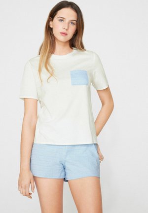 Junge Frau steht, trägt cremefarbenes T-Shirt mit blauem Brusttasche und hellblauen Shorts vor einfärbigem Hintergrund.