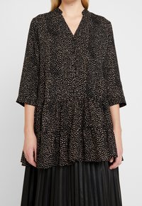 Czarna tunika z różowymi kropkami, dekoltem w kształcie v, rękawami o długości trzy czwarte, falbaną u dołu, wykonana z lekkiego materiału. Guziki z przodu.