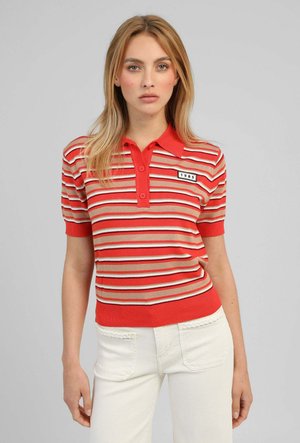 Femme portant un polo à manches courtes rayé rouge, beige et blanc avec un écusson "1985", assorti à un pantalon blanc sur fond gris.