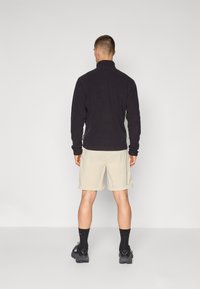 Musta fleece-takki korkealla kauluksella, yhdistettynä vaaleanbeigeihin shortseihin. Shortseissa on kääntöhelma, ja mallilla on mustat sukat ja kengät.