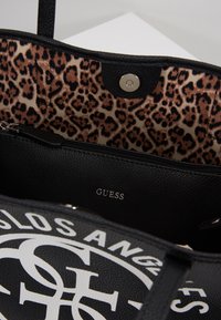 Czarna skórzana torebka z wnętrzem w panterkę, kieszenią na zamek i metalowym zatrzaskiem, z widocznym białym logo na zewnątrz.