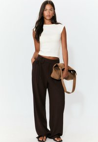 Bílý upnutý crop top, hnědé volné kalhoty s pružnou šňůrkou a kabelka z kamenného semiše. Holé nohy s jednoduchým designem.