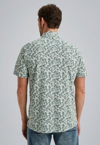 Camisa de manga corta en tela verde claro con un estampado de hojas. Presenta un cuello abotonado y un dobladillo curvo. Corte casual, vista posterior mostrada.