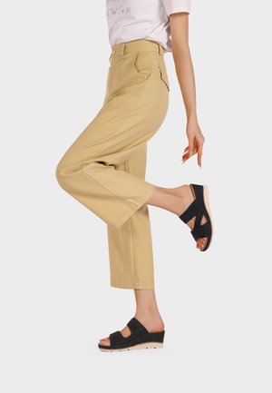 Pantaloni beige a gamba larga con tasche laterali, abbinati a sandali neri con zeppa, dotati di una tracolla testurizzata e una suola chiara a contrasto.