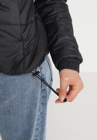 Sort puffjakke med en tekstureret overflade, der har elastiske ærmer og justering med snøre i taljen, båret med blå jeans.