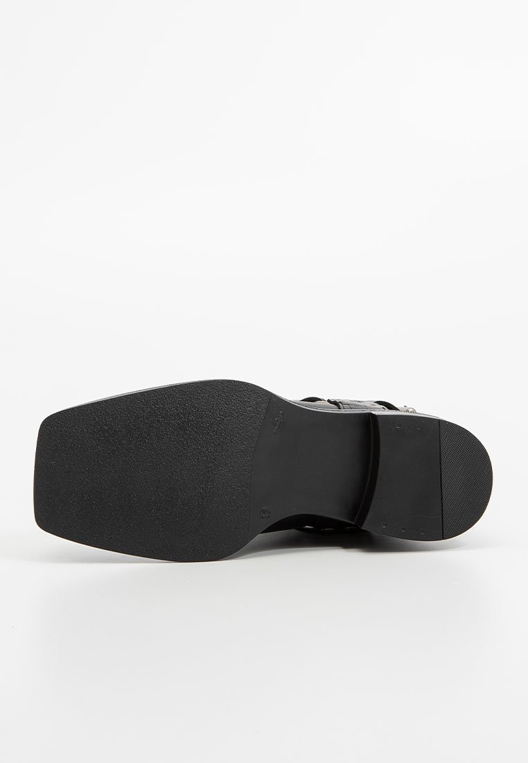 Sandale noire avec une semelle en caoutchouc lisse, de forme rectangulaire et surface texturée. Présente un design de courroie avec une esthétique minimaliste.