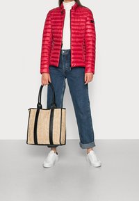 Rød puffjakke med høj krave og lynlås, parret med mørkeblå jeans. Modellen holder en strikket beige taske med sorte stropper.