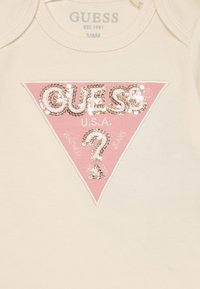 Bela otroška bodi z roza trikotnim logotipom z bleščicami, ki prikazuje "GUESS" in "ZDA" v kovinski niti, s poudarkom v obliki vprašaja.