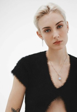 Top de punto negro y esponjoso con mangas cortas; collar de cadena plateada con colgante redondo de opalescente; cabello corto; maquillaje mínimo; fondo neutro.