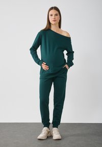 Laiša off-shoulder krekls, kas kombinēts ar saskaņotām treniņbiksēm, abi izgatavoti no mīkstas auduma. Apavi ietver gaismas krāsas sporta apavus.