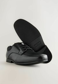 Zapatos de vestir de cuero negro con cordones y una suela texturizada, posicionados con un zapato ligeramente inclinado para resaltar el diseño y la marca.