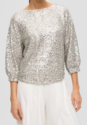 Femme portant un haut à manches longues en sequins argentés avec un col rond, associé à un pantalon blanc taille haute à jambes larges.