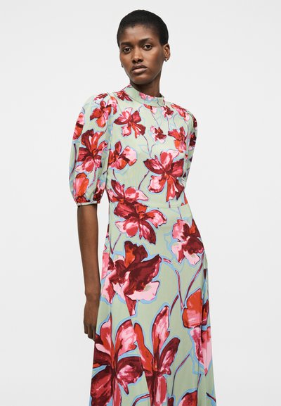 Γυναίκα που φοράει φόρεμα μέντας με μεγάλα κόκκινα και ροζ λουλουδάτα σχέδια, φουσκωτά κοντά μανίκια και ψηλό γιακά, στέκεται μπροστά σε λευκό φόντο.