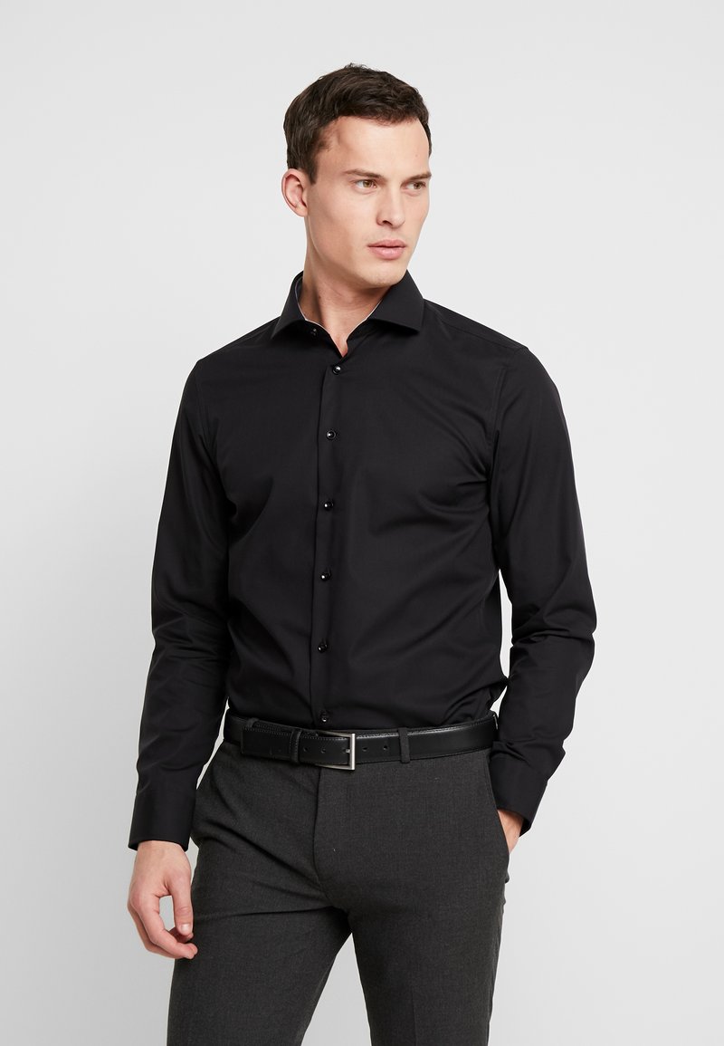 Homme portant une chemise noire ajustée boutonnée, rentrée dans un pantalon gris foncé avec une ceinture noire, debout sur un fond blanc uni.