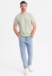 Lichtgroen gespikkeld T-shirt met een rond logo op de borst, gecombineerd met lichtblauwe rechte jeans en witte sneakers.