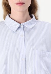 Camisa de botão listrada azul clara com colarinho clássico, um bolso no peito e tecido texturizado. Botões brancos complementam o design.