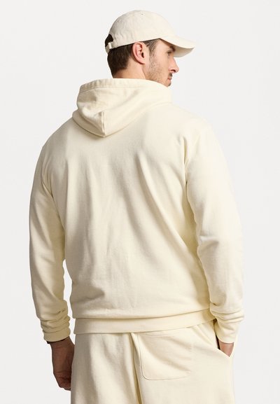Krämfärgad hoodie med känguruficka, mjuk textur och dragsko på huvan, bärs med matchande krämfärgade byxor och en keps.