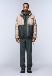 Gevoerde jas in beige en donkergroen, met een oranje rits, een oversized capuchon en elastische manchetten, gedragen over een donkergroene hoodie en broek.