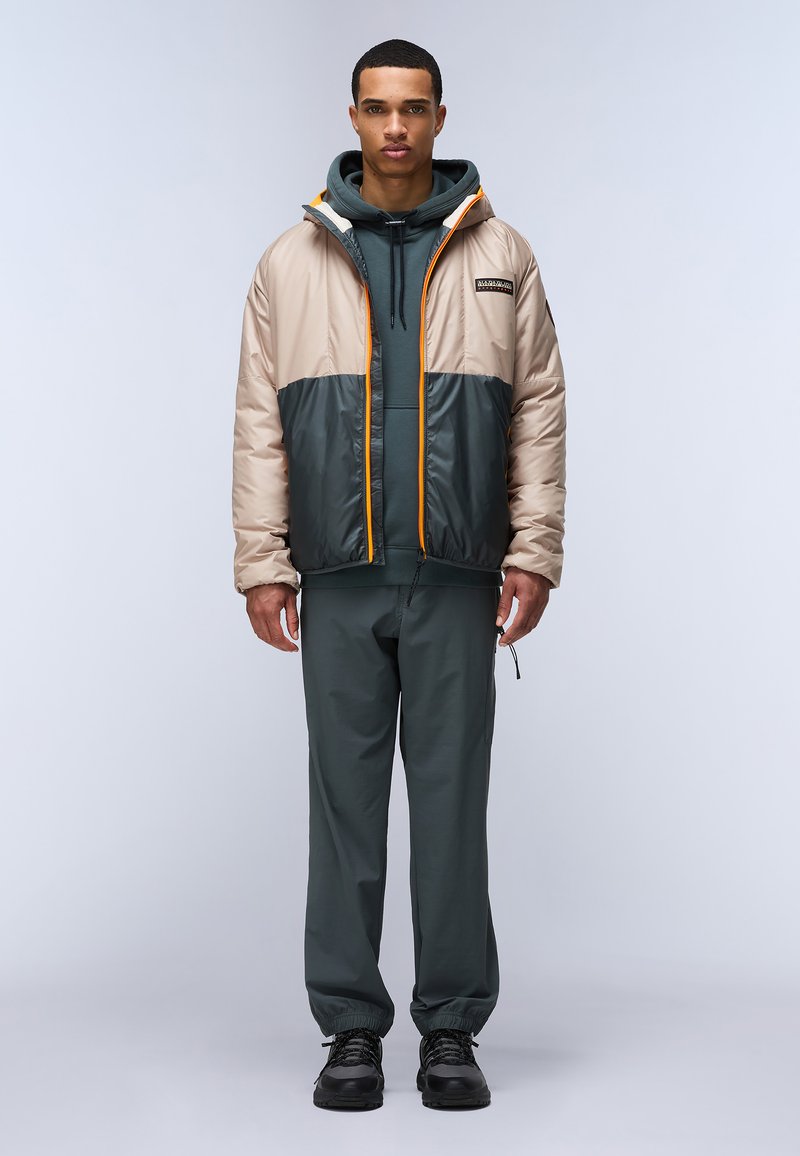 Chaqueta acolchada en beige y verde oscuro, con un cierre naranja, capucha extragrande y puños elásticos, llevada sobre una sudadera y pantalones de color verde oscuro.