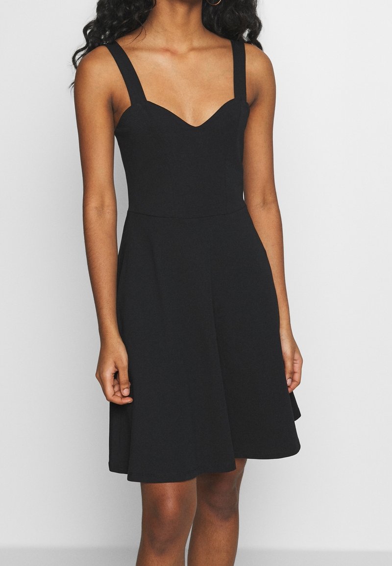Femme portant une robe noire sans manches mi-longue avec de larges bretelles et un corsage ajusté, debout devant un fond clair uni.