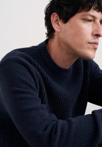 Jūras spēku zila adīta džempera ar teksturētu vafeļu rakstu un ribotu apkakli, izceļot biezu audumu un pieguļošas piedurknes.