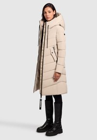 Vrouw die staat in een lange beige gewatteerde jas met grote capuchon, zwarte leggings en kniehoge zwarte chunky laarzen, iets naar links gekeerd.