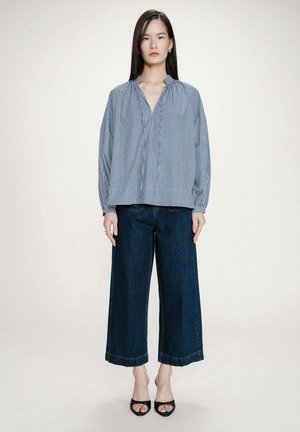 Femme portant un chemisier ample à carreaux bleus et blancs avec un jean large bleu foncé et des talons noirs ouverts, debout devant un fond uni.