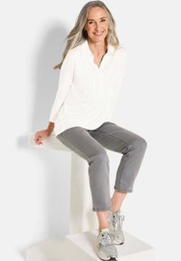 Top bianco a maniche lunghe con pieghe sottili, abbinato a jeans grigi a vita alta e scarpe da ginnastica argentate, seduto su un piedistallo di colore chiaro.