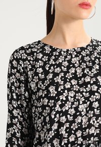 Musta kukallinen pusero, jossa on pyöreä pääntie, pitkät hihat ja valkoisten kukkien kuvio, jossa on hienovaraisia vaaleanpunaisia yksityiskohtia kevyessä kankaassa.