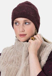 Bonnet en tricot bordeaux avec un motif texturé, associé à un gilet en polaire crème à fermeture éclair. Le modèle porte un grand anneau et des boucles d'oreilles pendantes.