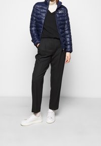 Person som bär en marinblå vadderad jacka, svarta byxor, vit polotröja, svart väst, vita strumpor och vita sneakers, stående med händerna i fickorna.