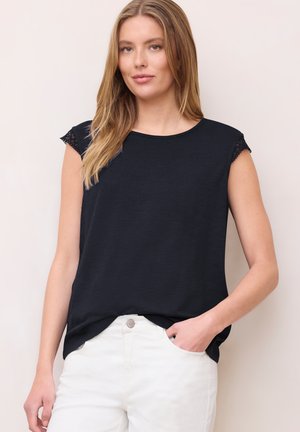 Kvinde iført en ærmeløs sort top med blondekant og hvide bukser, stående med den ene hånd i lommen mod en lys baggrund.