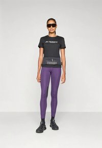 Kvinde iført sort Terrex t-shirt og solbriller, lilla leggings, sorte sneakers og en sort Terrex talje taske, stående foran en hvid baggrund.