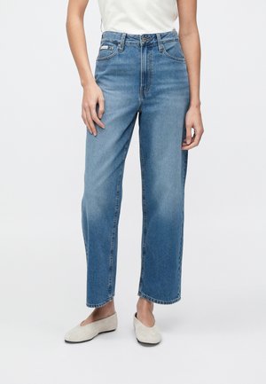 Frau trägt blaue High-Waist-Jeans mit weitem Bein, weiße Slip-on-Schuhe und ein weißes ärmelloses Oberteil, steht vor einem einfarbigen Hintergrund.